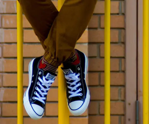 Person in khaki pants with black converse sitting on yellow fence in front of school.
