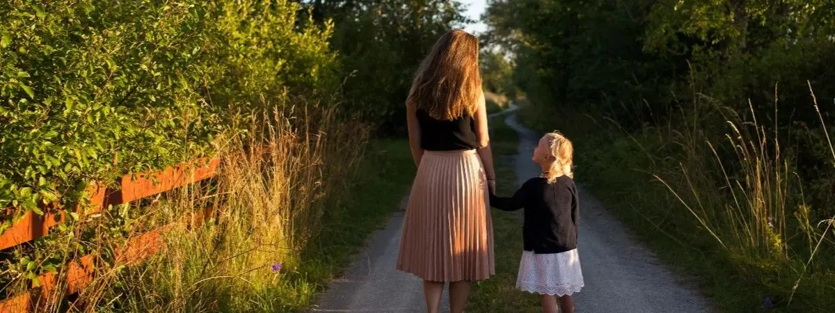adult holding hands with child walking on road surrounded by grass and tree