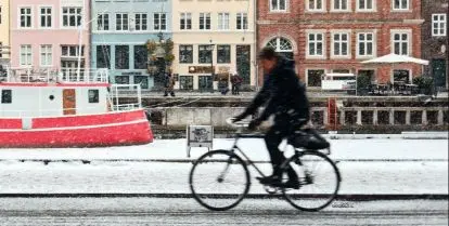 person riding bicycle on snowy street with boat and colorful buildings behind
