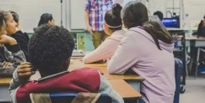 young students sitting around table looking at person standing at the front of classroom